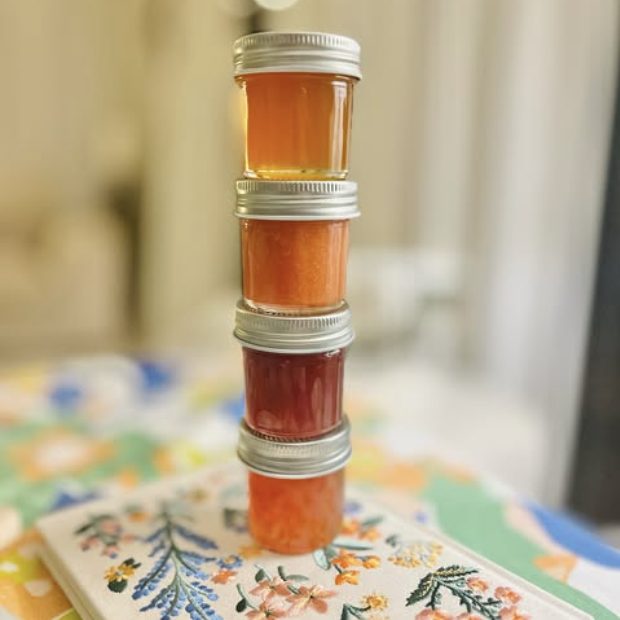 Four small jars stacked on an embroidered cloth: honey, marmalade, jam